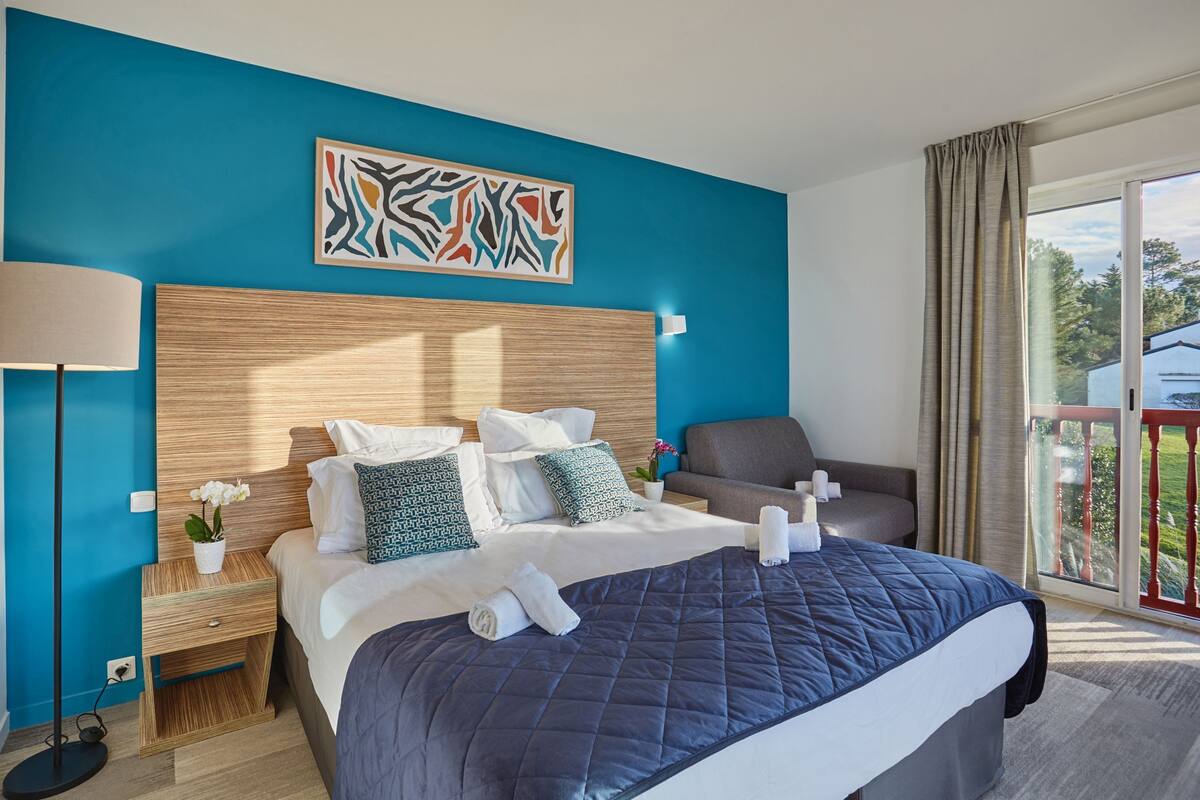 Ein Hotelzimmer mit einem großen Bett, einem hölzernen Kopfteil, einer blauen Akzentwand, einer Stehlampe und Blick ins Freie.