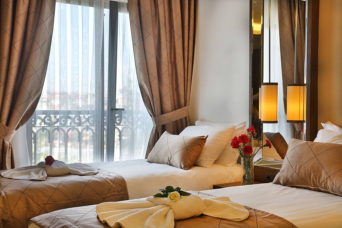 A hotel room with two beds, a large window with curtains, a vase with flowers, and a nightstand with lamps.