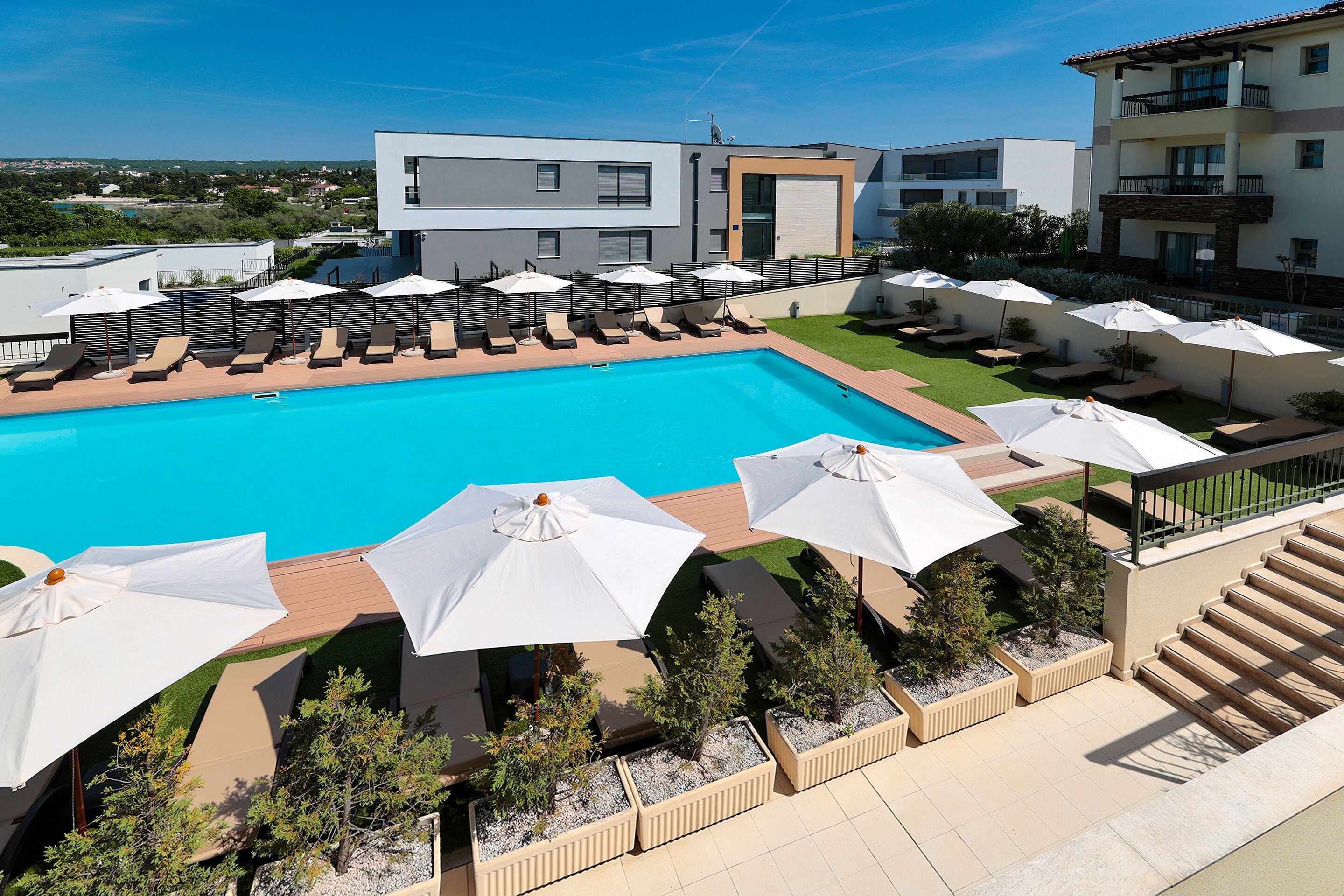 Un área de piscina moderna con sillones y sombrillas, rodeada de edificios con balcones y una vista despejada del paisaje.