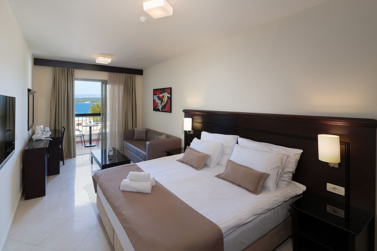 Habitación de hotel con cama, lámparas de noche, escritorio con silla, televisor y vista al mar desde el balcón.