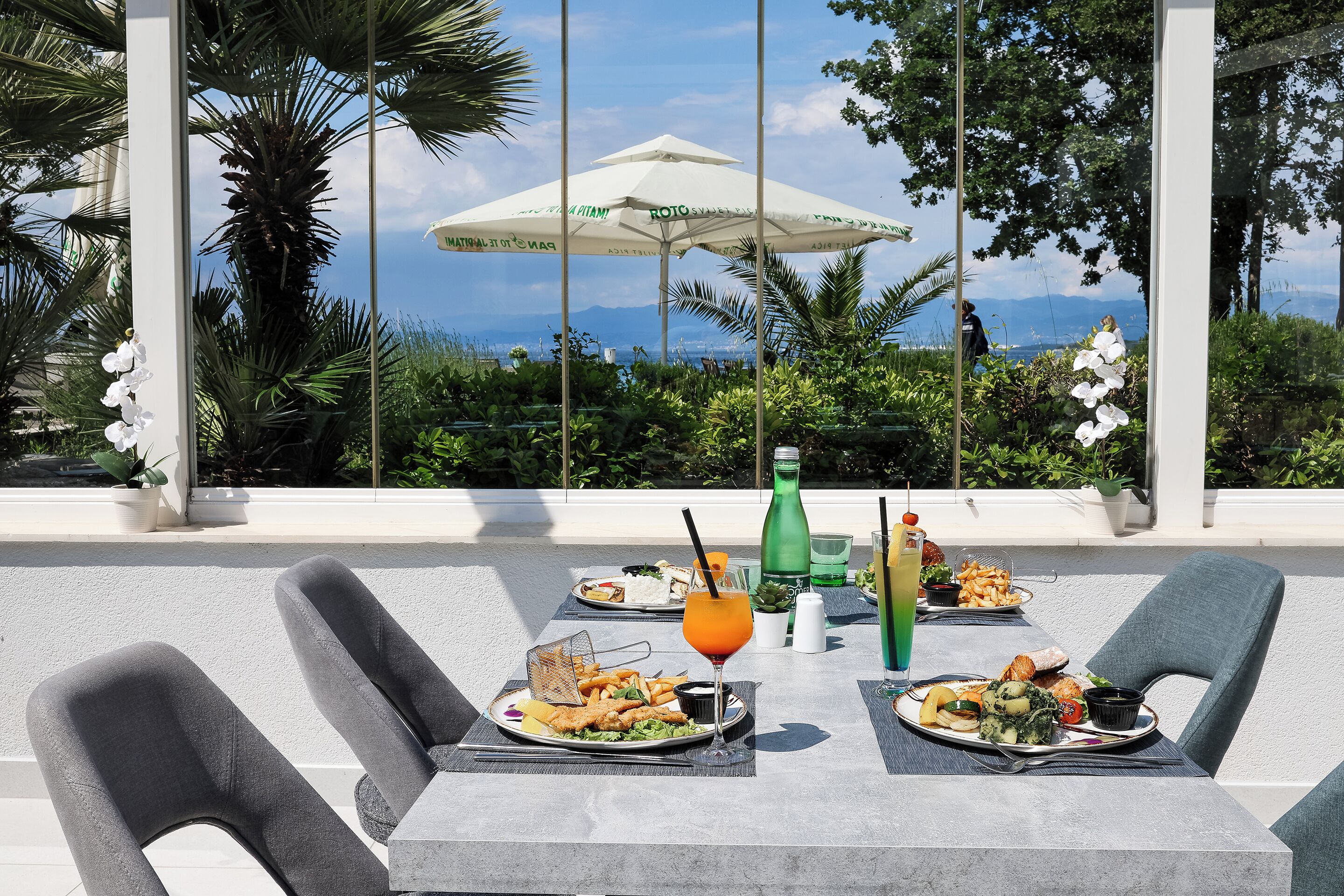 An outdoor dining area with two tables, chairs, a white umbrella, and a view of the ocean.