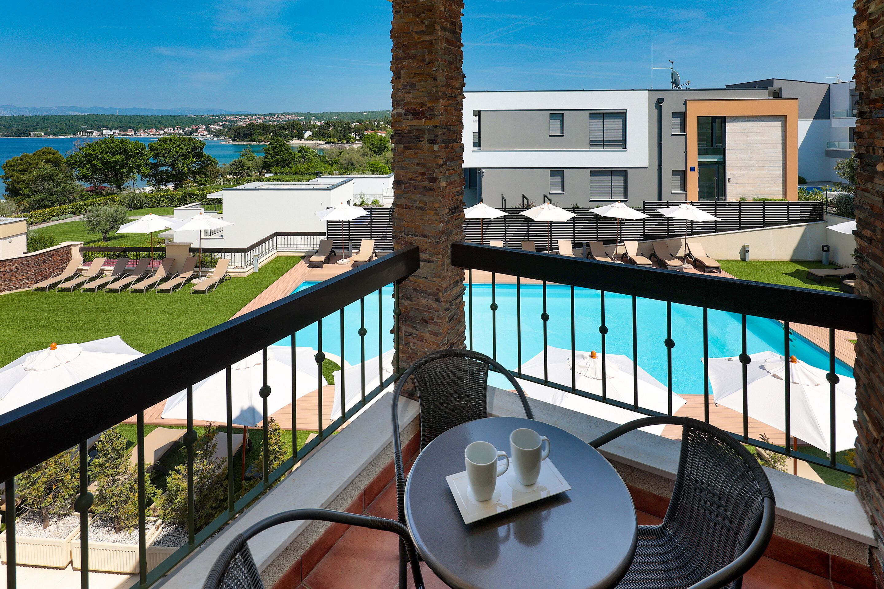 Ein Balkon mit Pool, Liegestühlen und Sonnenschirmen, mit Blick auf ein modernes Gebäude und die Küste.