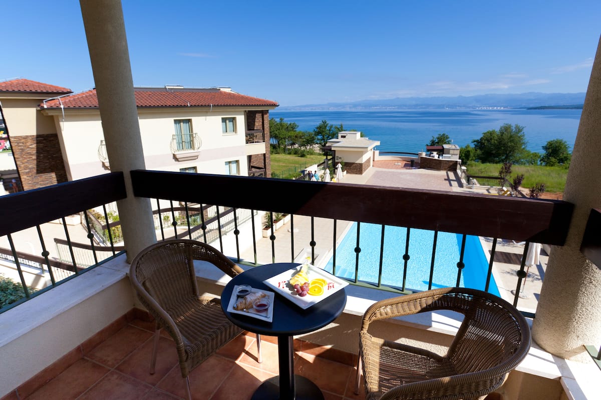Un balcón con sillas de mimbre, una mesita con una bandeja de fruta y una bebida, con vistas a una piscina y al mar y edificios.