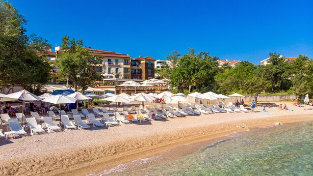 A beach with sun loungers and umbrellas, clear water, and buildings in the background. - Blue Waves Resort (Malinska-Dubasnica)