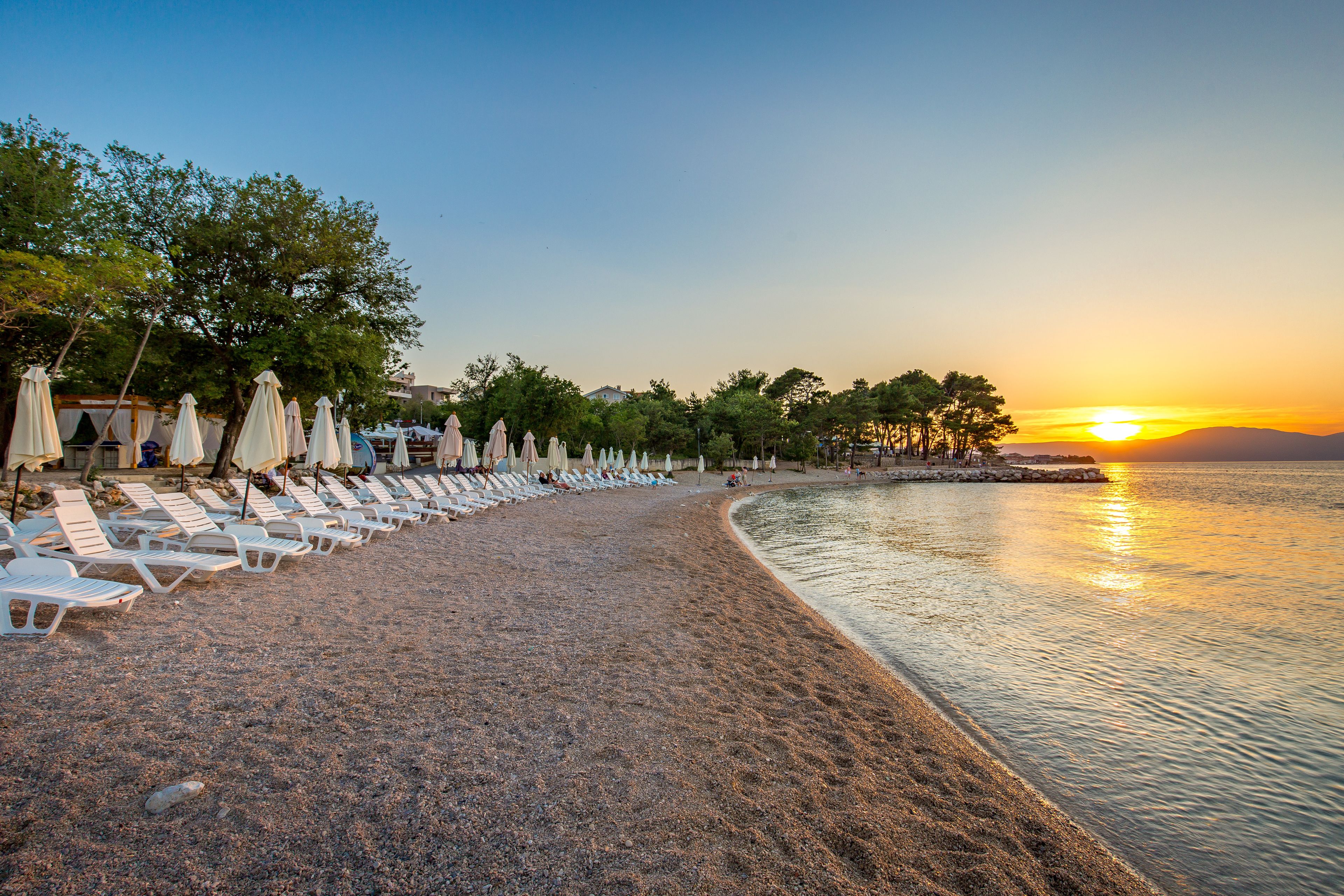 A beach with rows of white lounge chairs and umbrellas, a calm sea, and a sunset in the background.