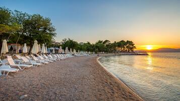 A beach with rows of white lounge chairs and umbrellas, a calm sea, and a sunset in the background.