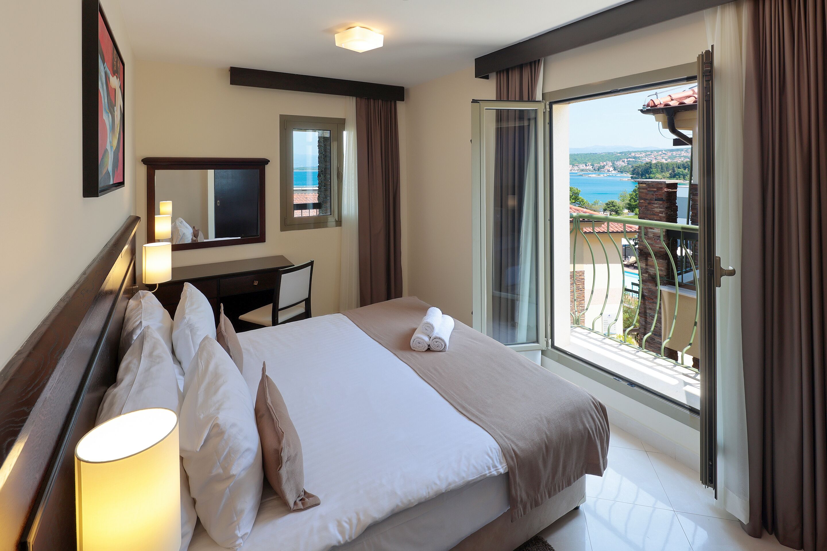 A hotel room with a bed, a desk, a chair, a lamp, a mirror, and a balcony with a view of the sea.