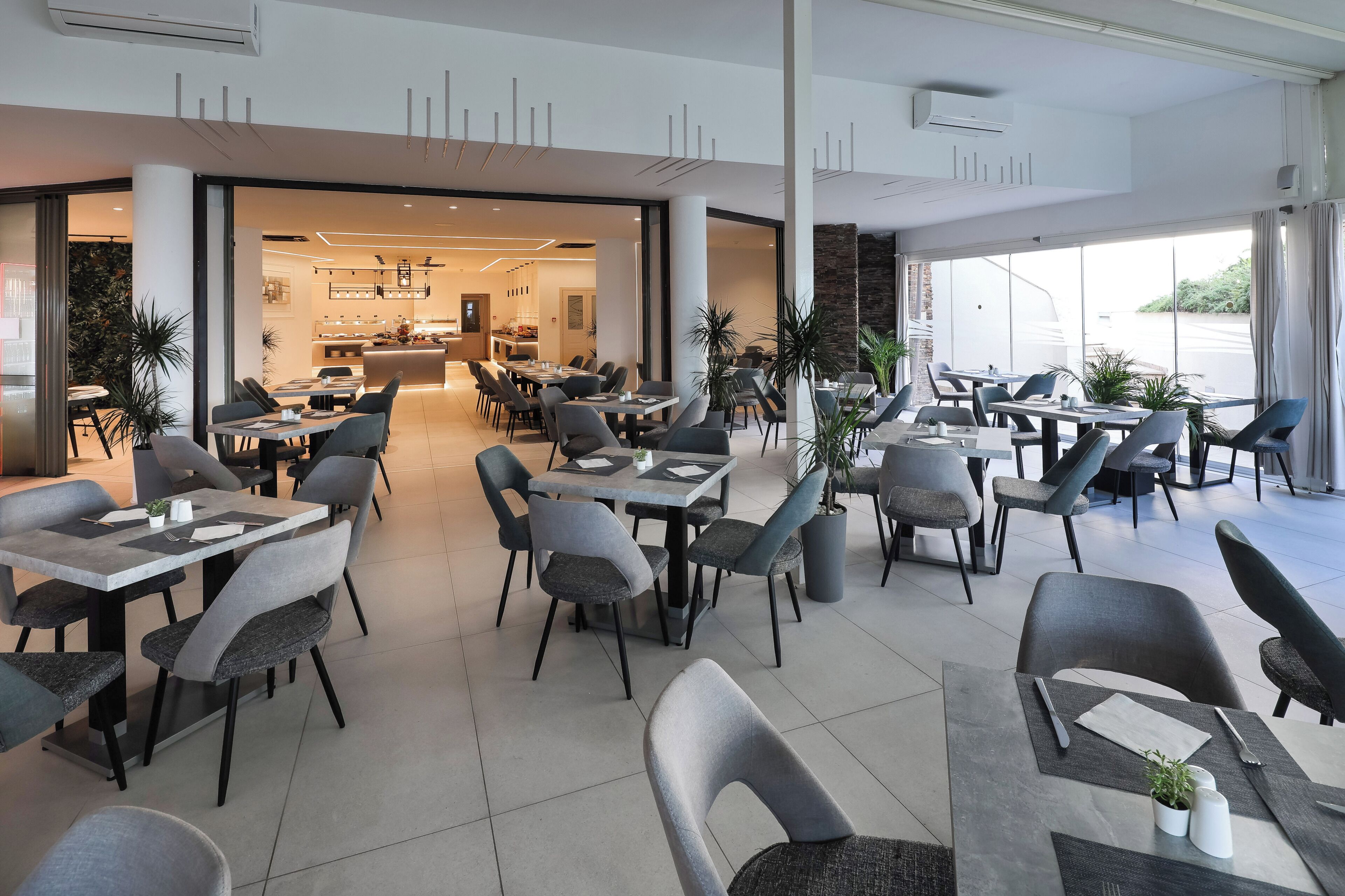 A modern dining area with tables and chairs, a kitchen in the background, and large windows.