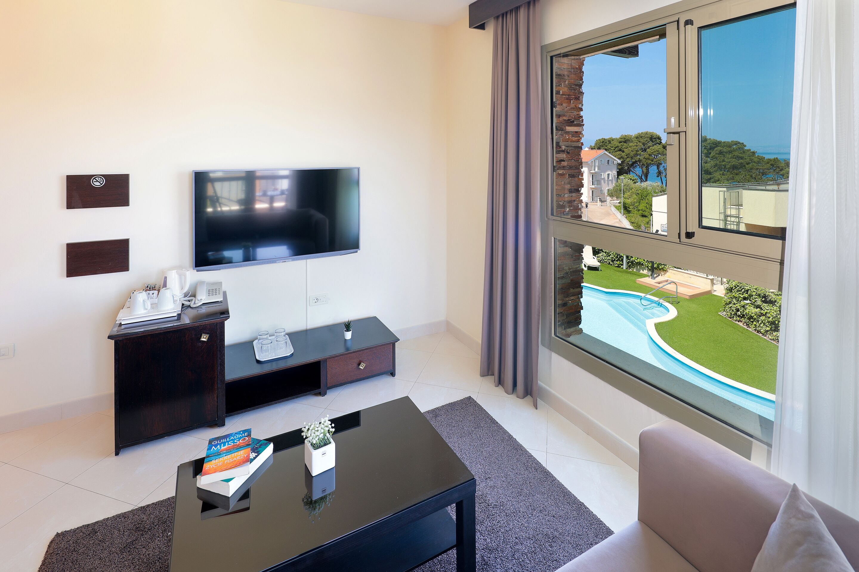 A modern living room with a flat-screen TV, dark wooden furniture, a coffee table with books, and a large window with a view of a swimming pool and greenery.