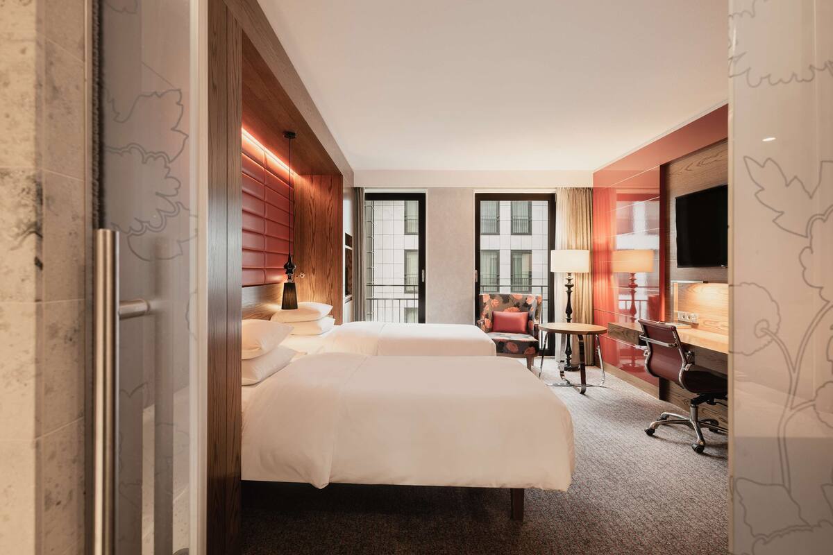 A hotel room with two beds, a desk, a chair, a TV, and a window.