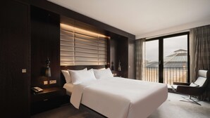 A modern hotel room with a large bed, a nightstand, a desk, a chair, and a view of the city through the window. - Hilton The Hague (The Hague)