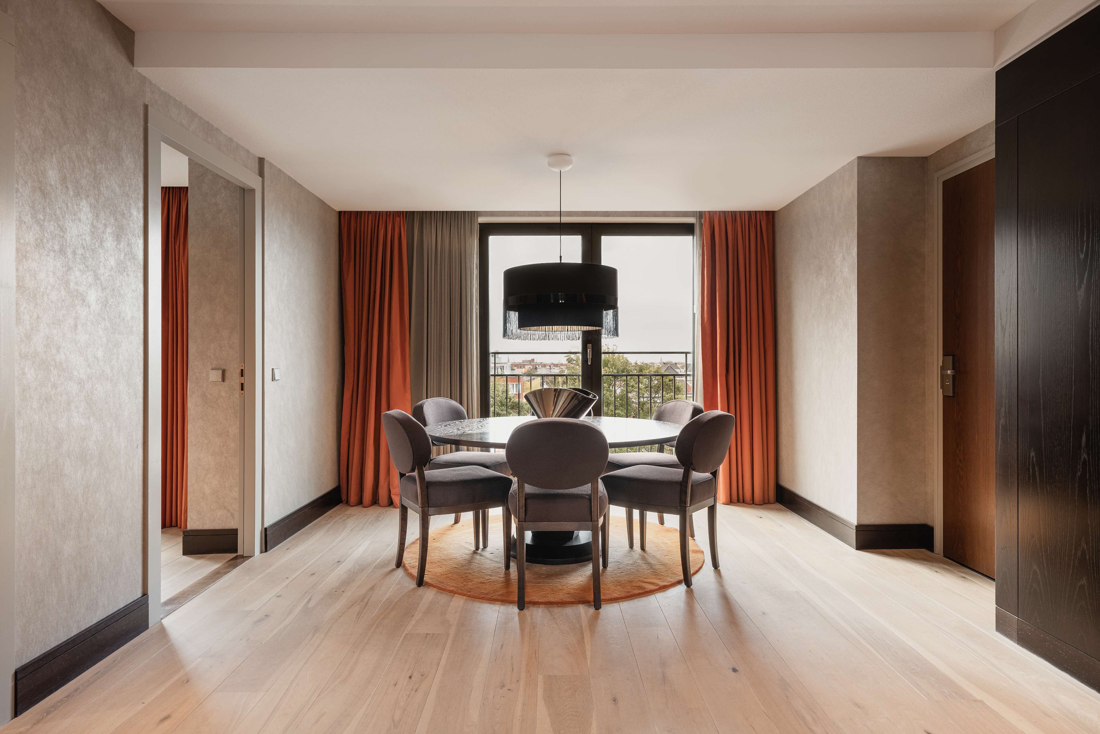 A modern dining area with a round table and six chairs, a large window with curtains, and a view of the city.
