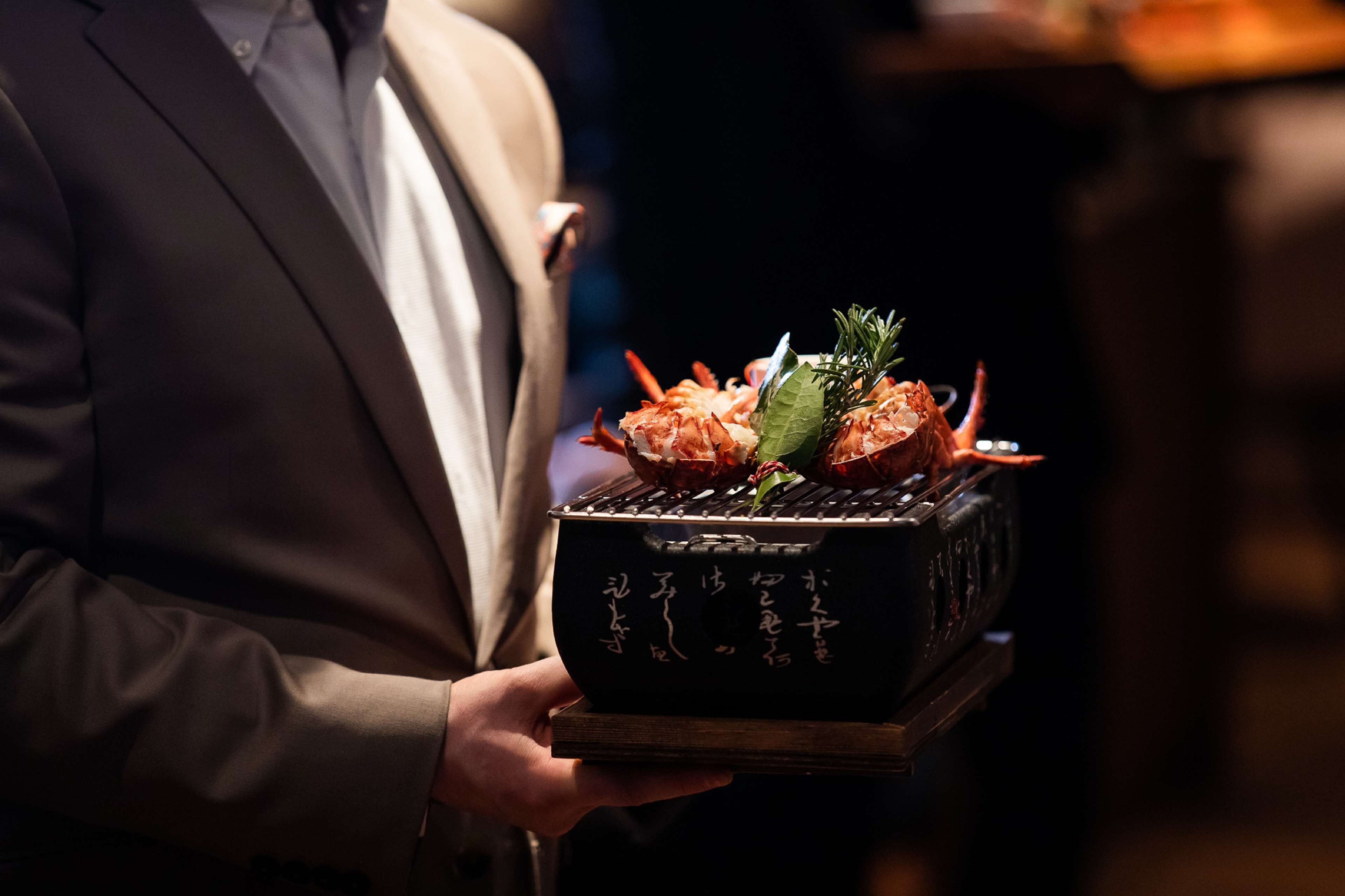 A person holding a tray with grilled shrimp and a sprig of rosemary.