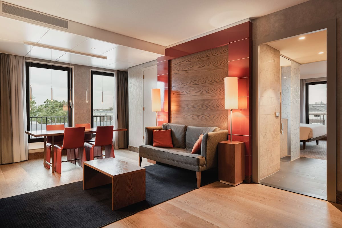 A modern hotel room with a sofa, dining table, chairs, and a large window offering a city view.