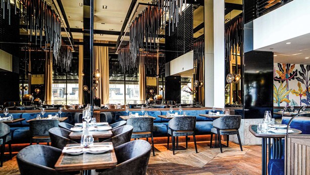 A modern restaurant interior with high ceilings, hanging light fixtures, and blue upholstered seating. - Hilton The Hague (The Hague)