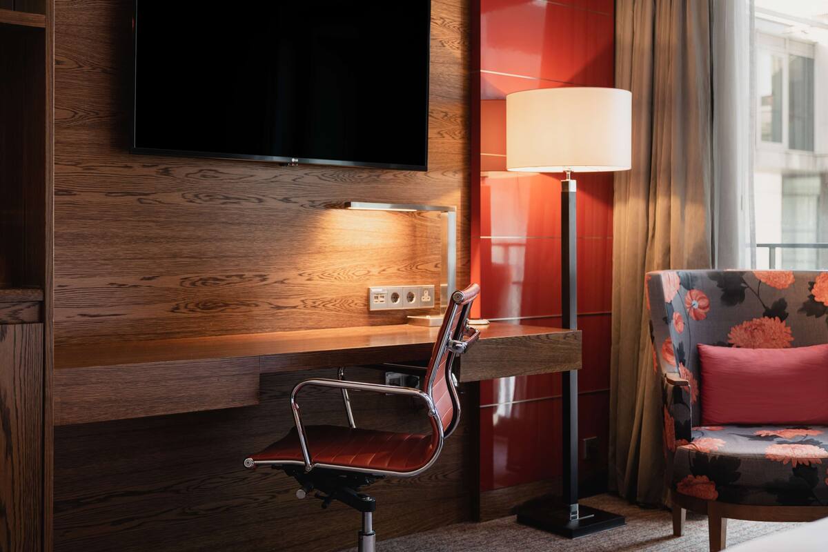 A modern hotel room with a wooden wall, a flat-screen TV, a red upholstered chair, and a floor lamp.