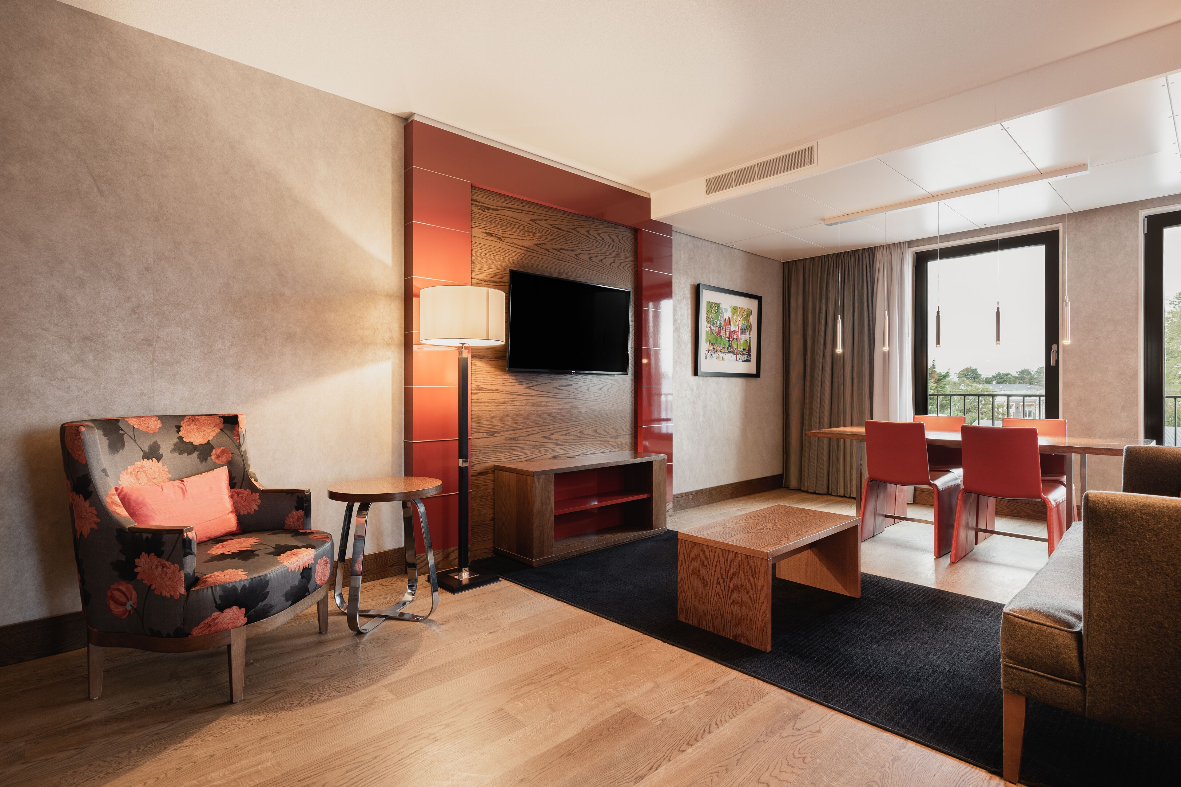 Una habitación de hotel moderna con suelo de madera, un sofá tapizado con motivos florales, una mesita auxiliar pequeña, un televisor de pantalla plana montado en una pared roja decorativa, una mesa de comedor con sillas rojas, y un ventanal con vistas a la ciudad.