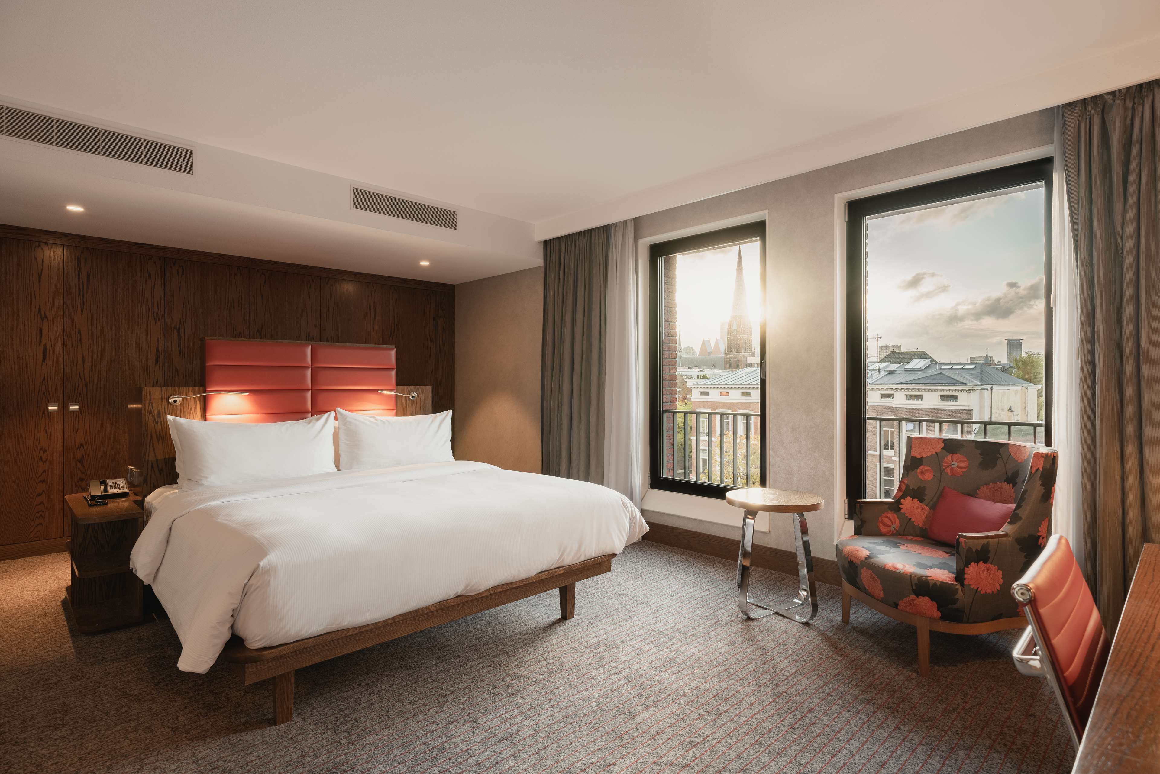 A hotel room with a large bed, a chair, a small table, and a view of the city through the window.
