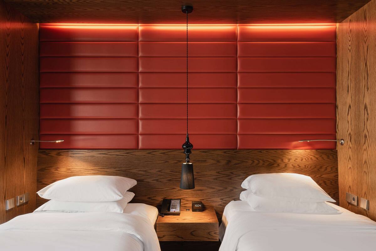 A hotel room with two beds, a wooden headboard, a bedside table with a lamp, and a clock.