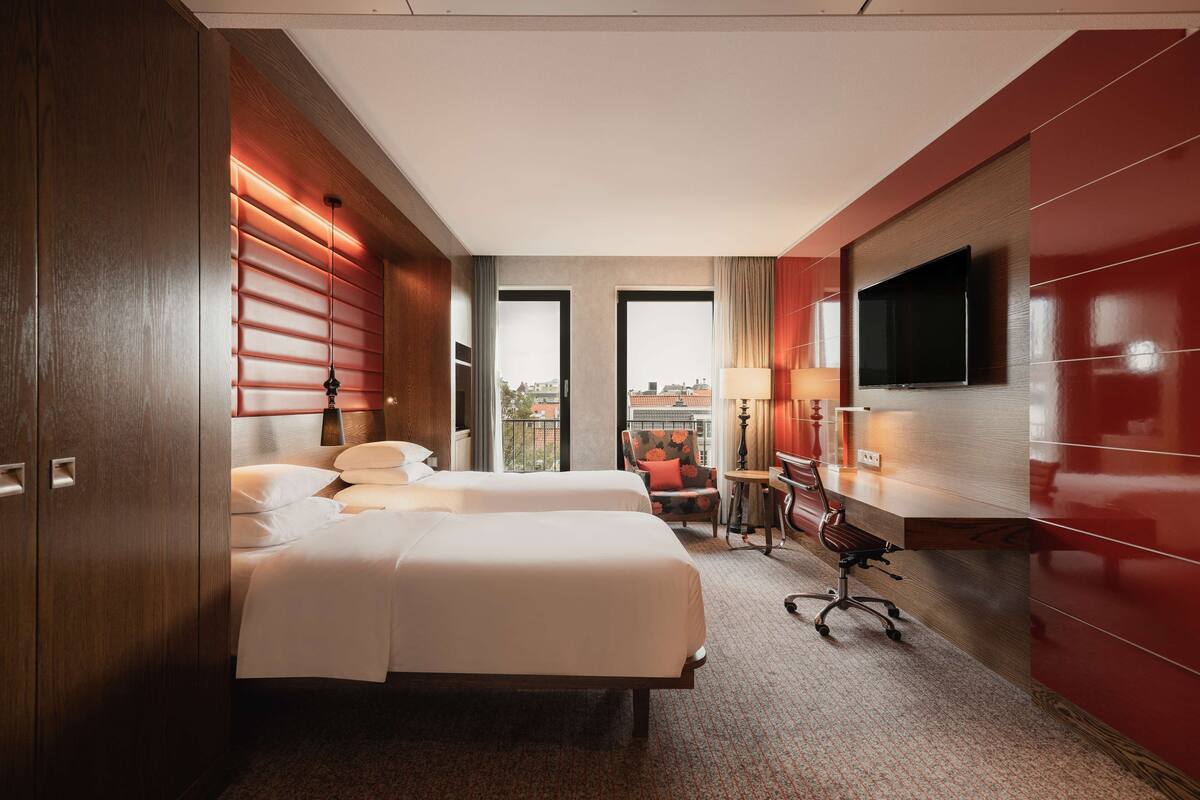 A hotel room with two beds, a desk, a chair, a TV, and a view of the city.