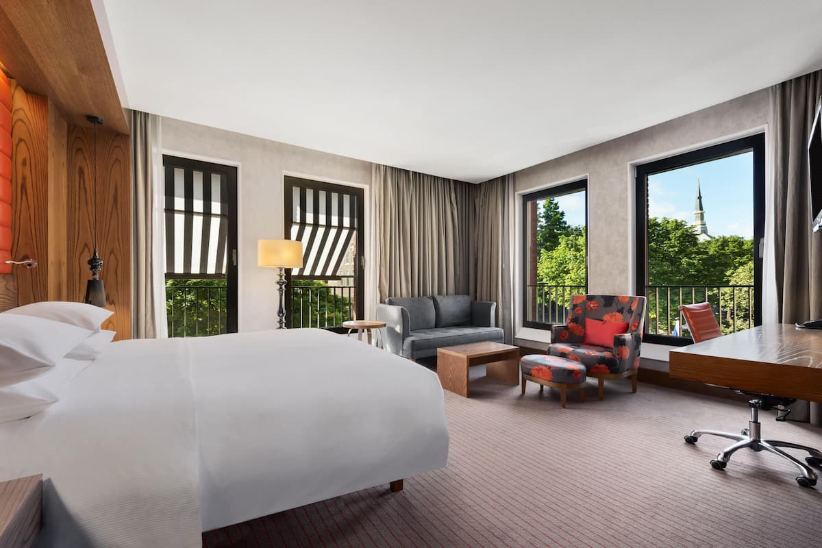 A hotel room with a bed, a sofa, a chair, a desk, and large windows offering a view of greenery and a building.