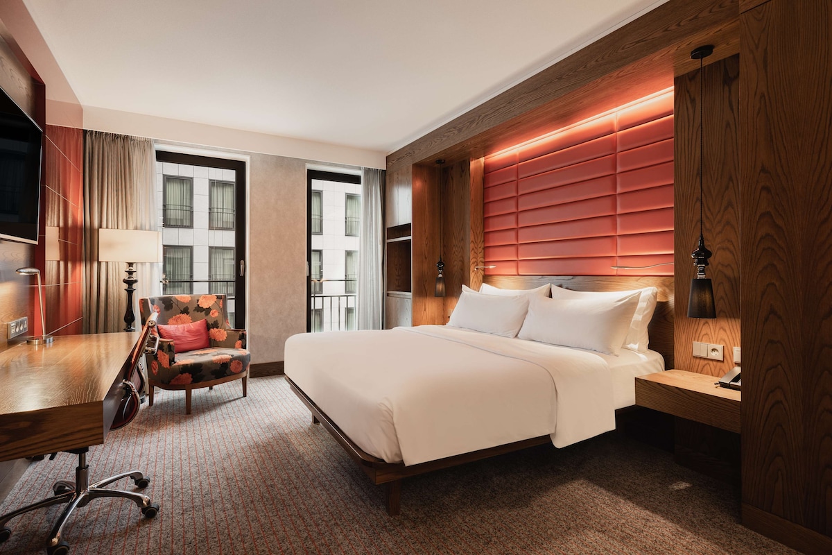 Habitación de hotel con una cama grande, un escritorio con silla, un televisor y una ventana con cortinas.