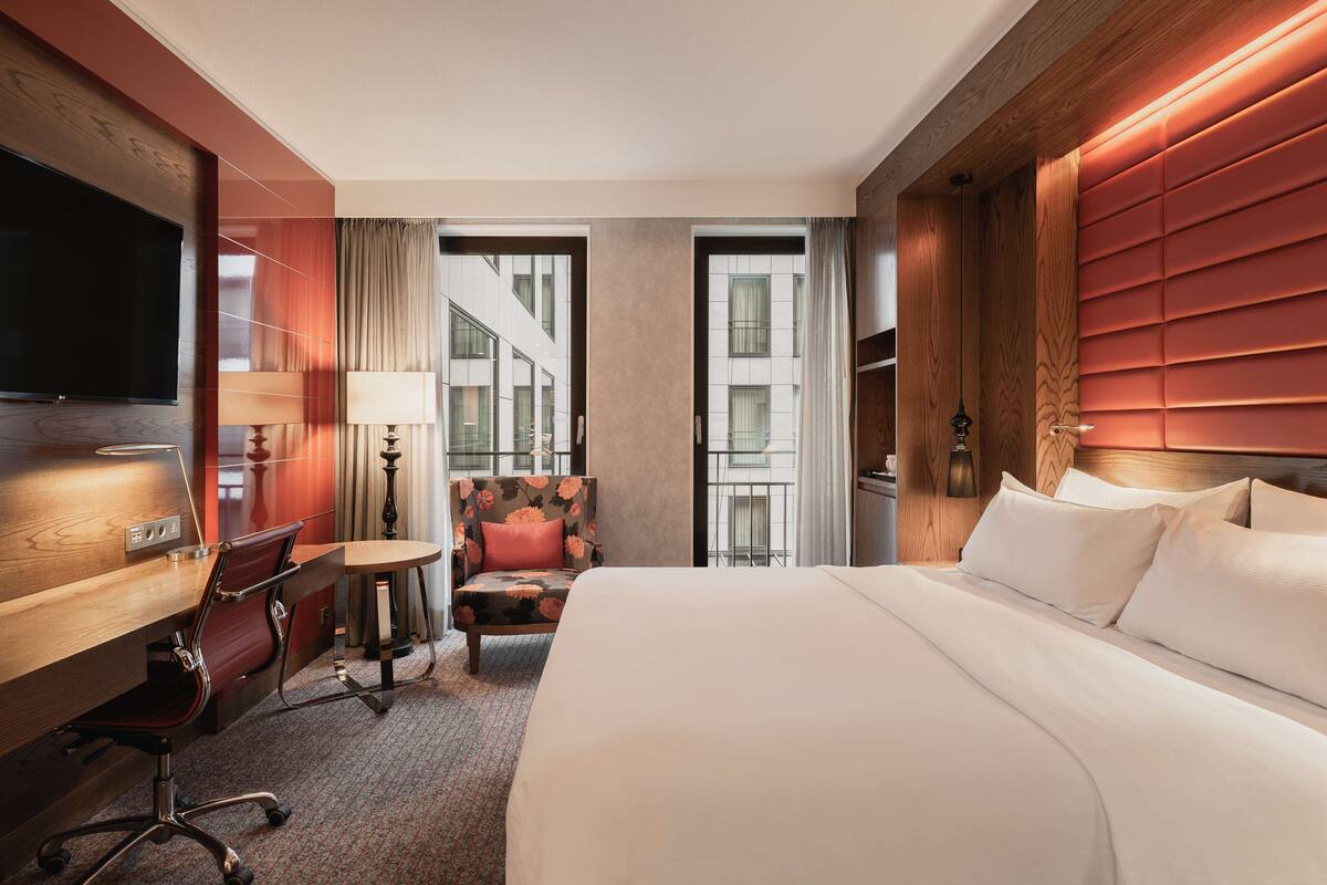 A hotel room with a large bed, a desk with a chair, a small table with a lamp, and a view of the city through large windows.