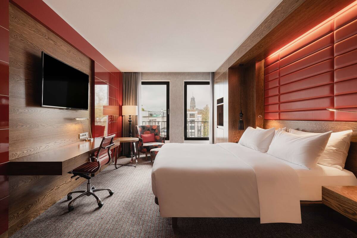 A hotel room with a large bed, a desk with a chair, a TV mounted on the wall, and a view of the city through the windows.