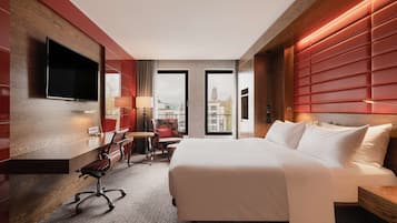 Een hotelkamer met een groot bed, een bureau met stoel, een aan de muur bevestigde televisie en uitzicht op de stad door de ramen.