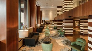 A spacious lobby with a modern design, featuring a central seating area with green chairs, a marble coffee table, and a large window providing a view of the outside. - Hilton The Hague (The Hague)