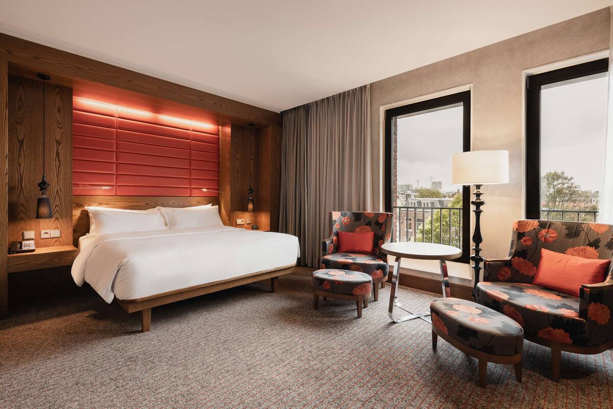 A hotel room with a large bed, a seating area with a table, and a view of the city through large windows.
