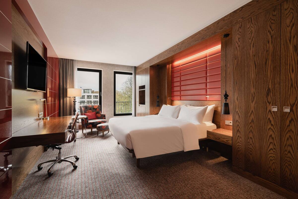 A modern hotel room with a large bed, a desk with a chair, a TV, and a view of the outside.