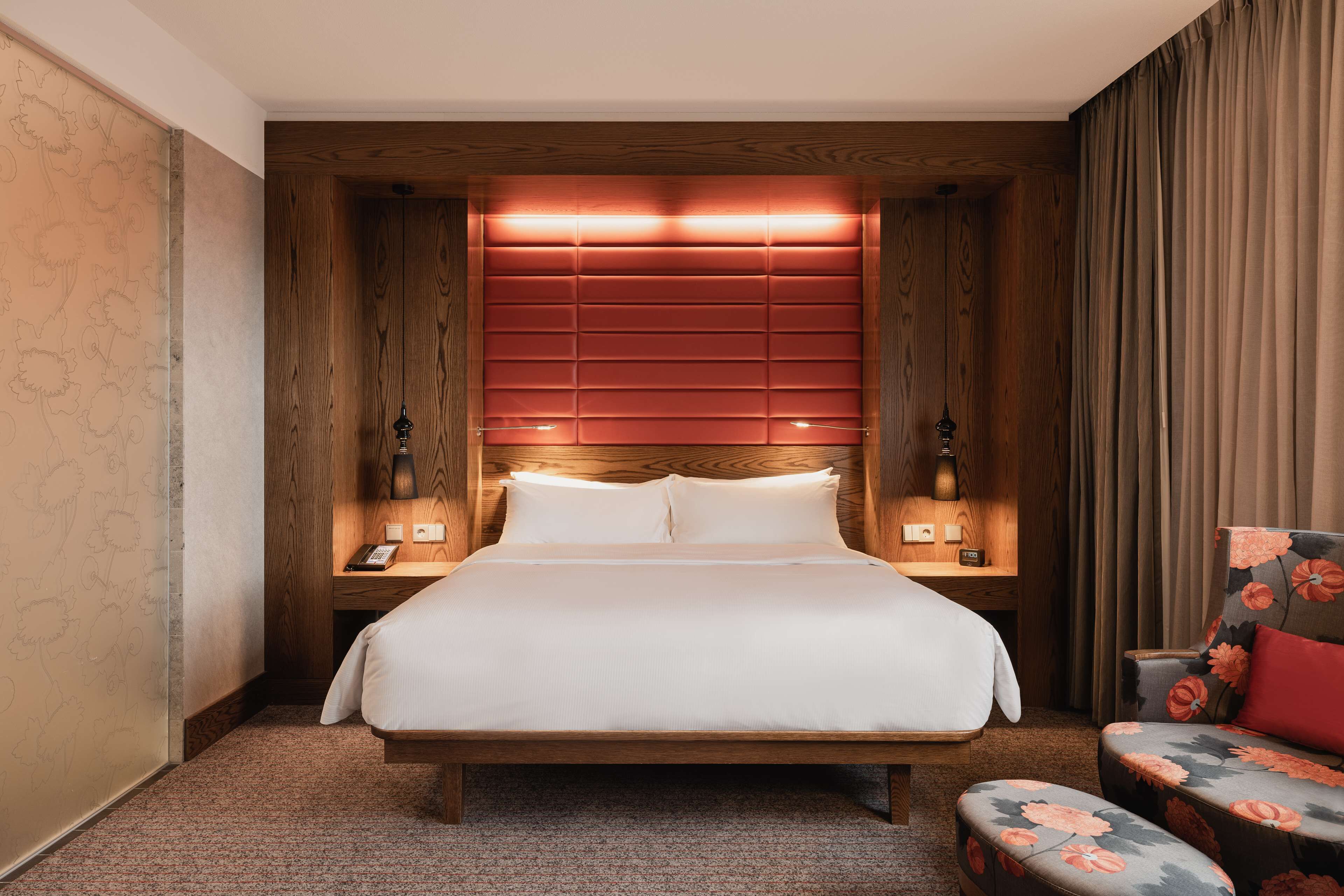 A hotel room with a wooden headboard, a large bed, a bedside table with a lamp, and a floral armchair.