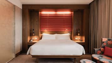 A hotel room with a wooden headboard, a large bed, a bedside table with a lamp, and a floral armchair.
