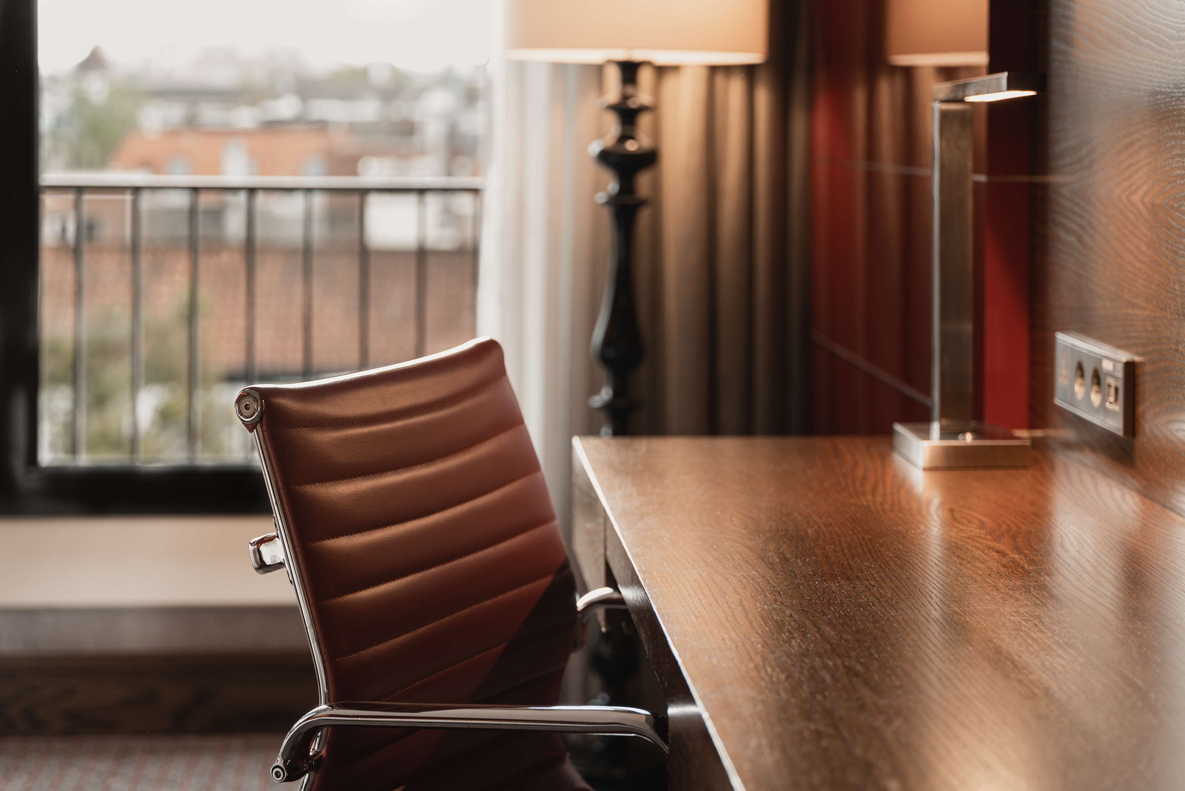 Une chambre d’hôtel avec une table en bois, un fauteuil en cuir, une lampe et une vue sur les immeubles par la fenêtre.