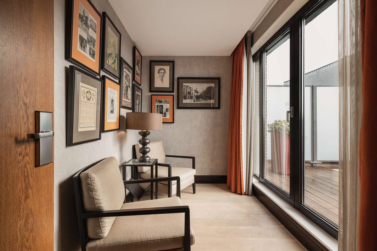 A hotel room with a large window, a sofa, a chair, and framed pictures on the wall.