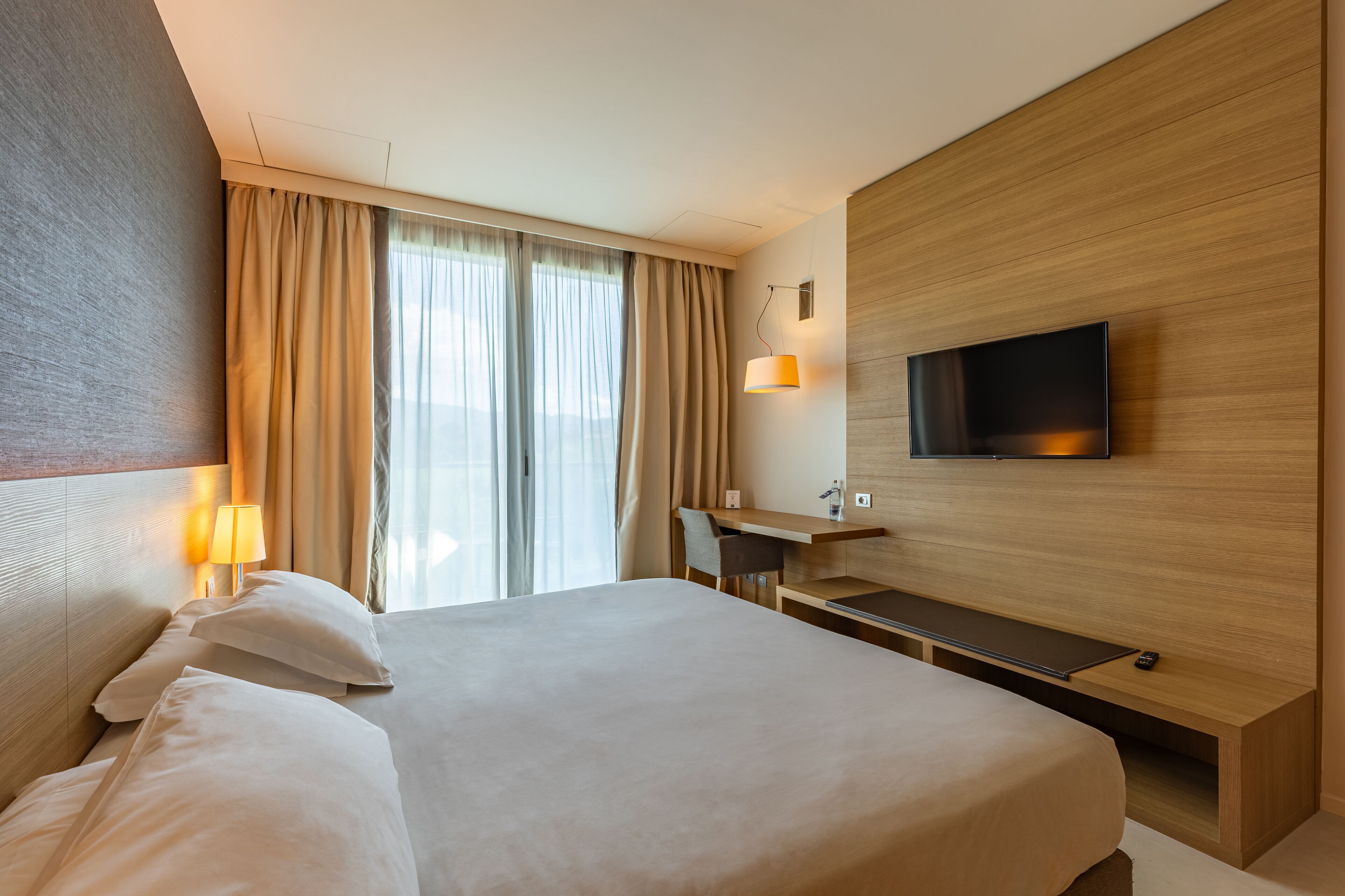 Ein modernes Hotelzimmer mit einem großen Bett, einem Schreibtisch, einem an der Wand montierten Fernseher und einem Fenster mit Vorhängen.