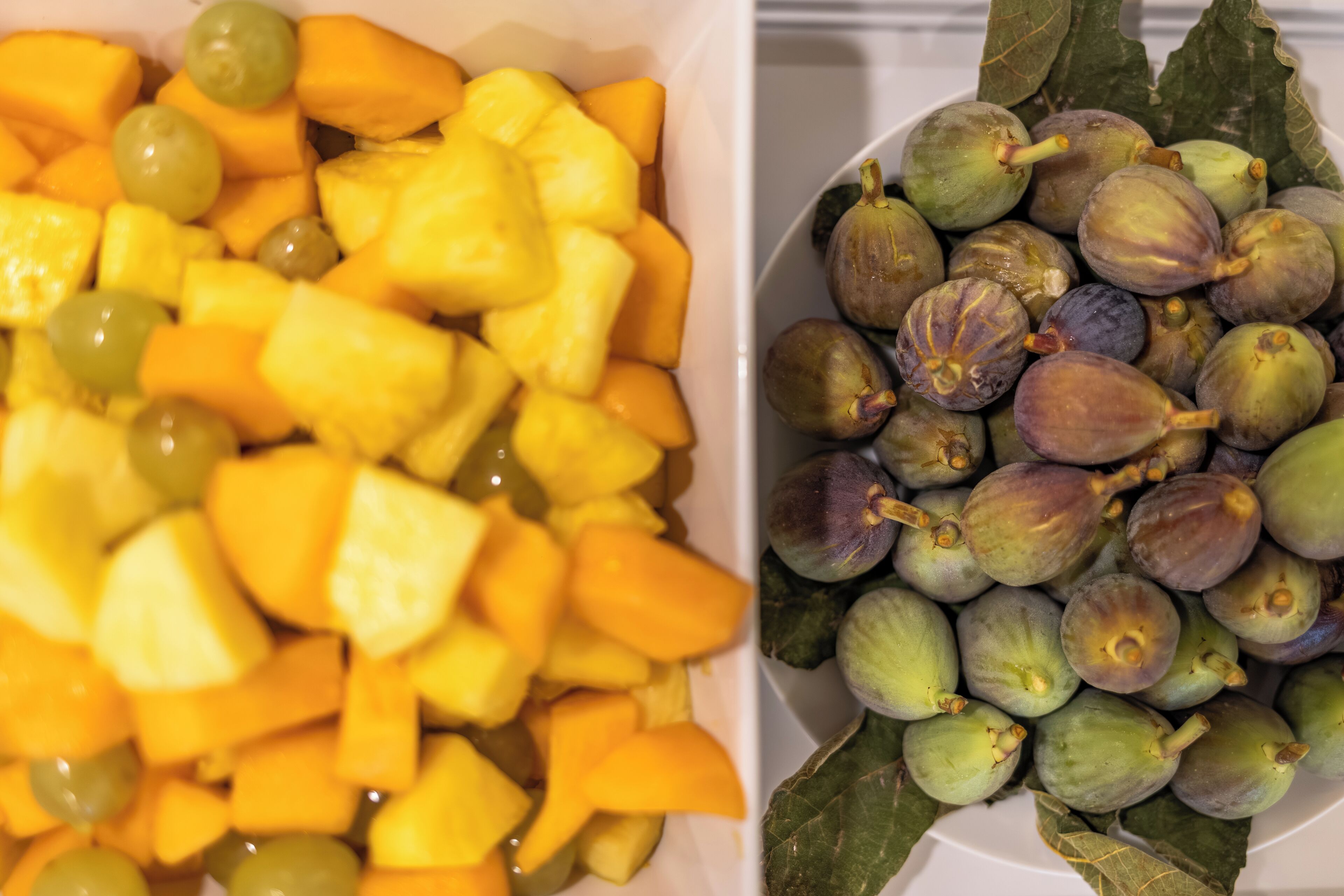 Two bowls of fruits, one with pineapple chunks and grapes, and the other with figs and leaves.