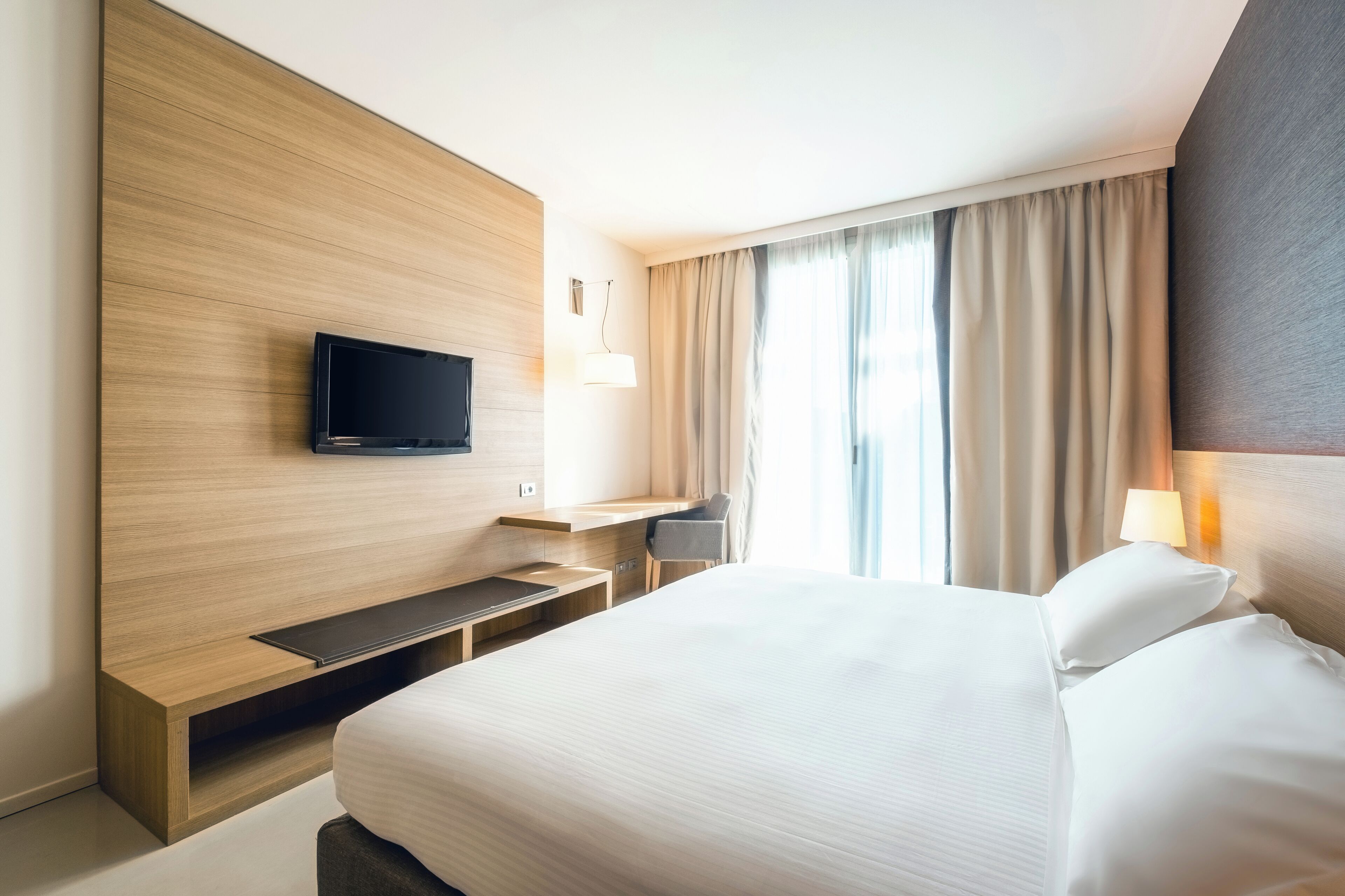 A modern hotel room with a large bed, a TV mounted on a wooden panel, a desk with a chair, and a window with curtains.