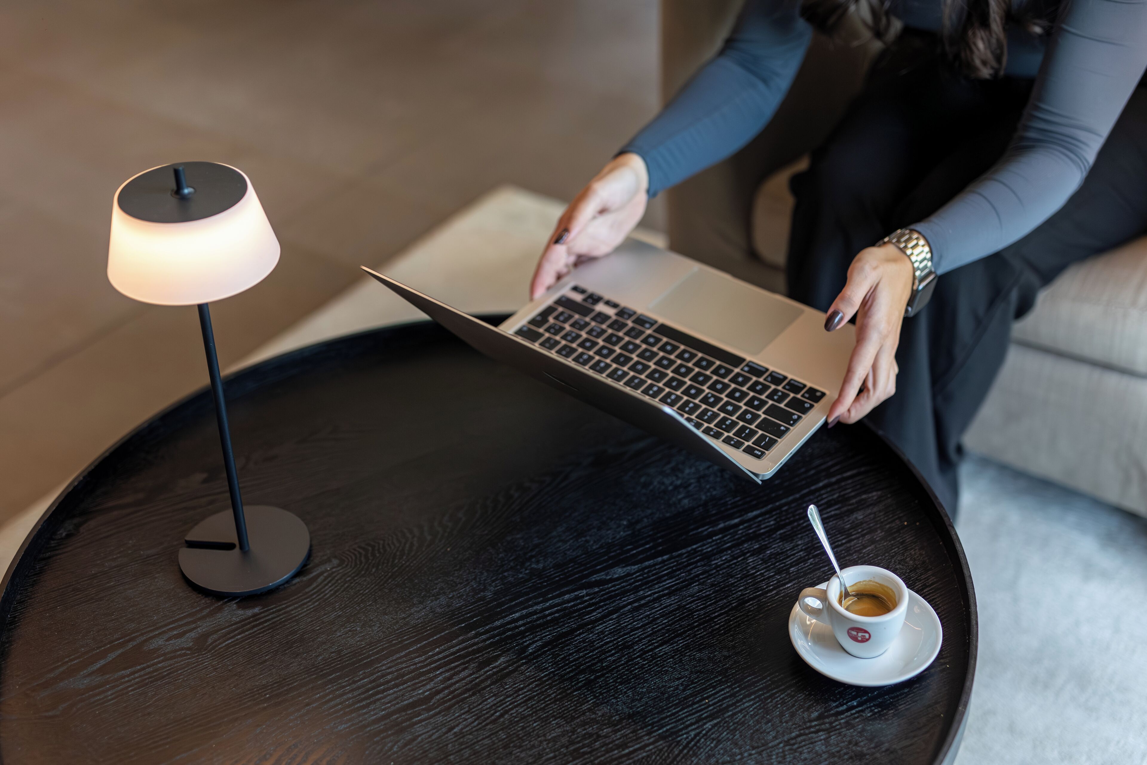 A laptop on a round black table with a white cup of coffee and a lamp beside it.