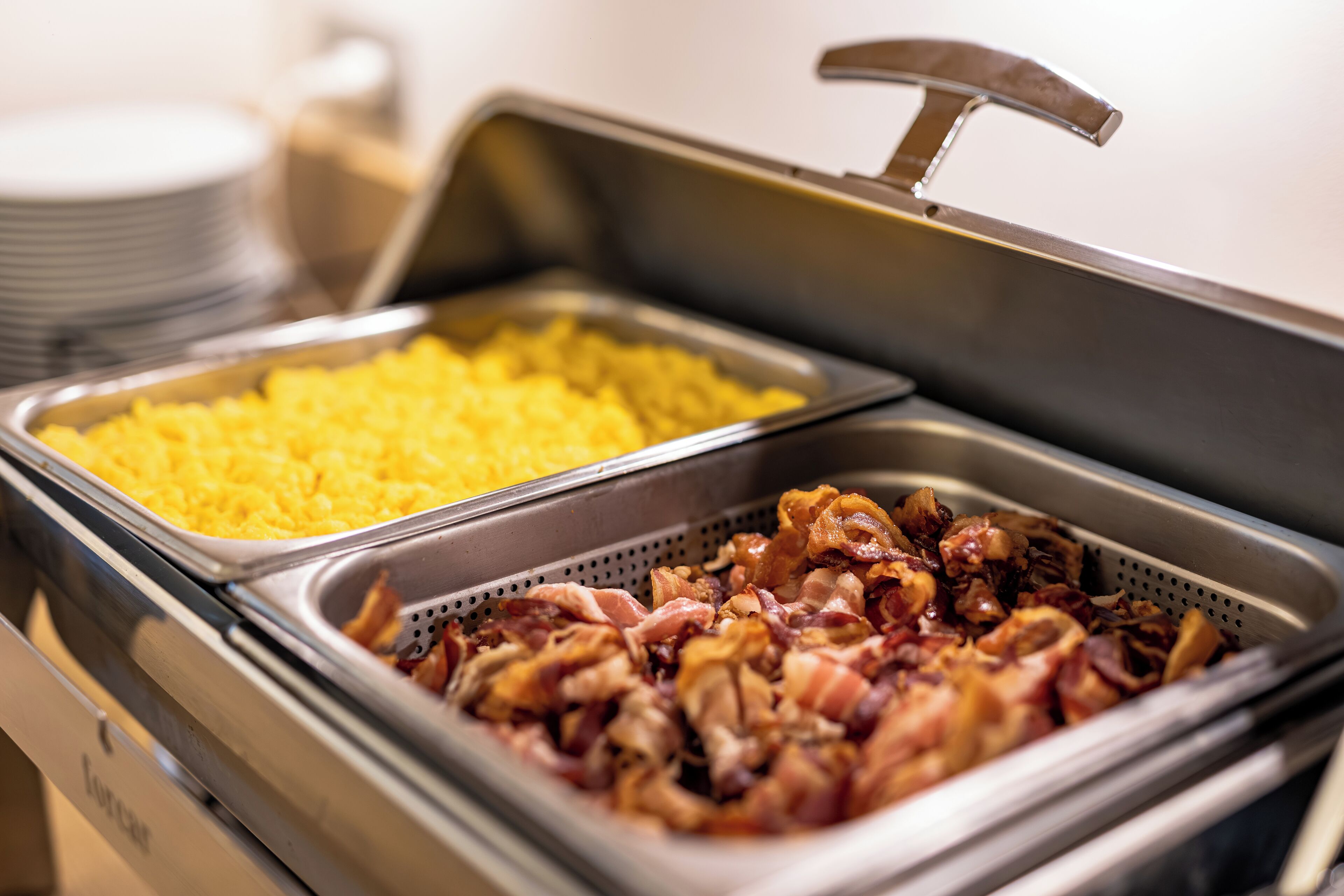 Un bufé con dos fuentes de comida: una con huevos revueltos y la otra con tocino.