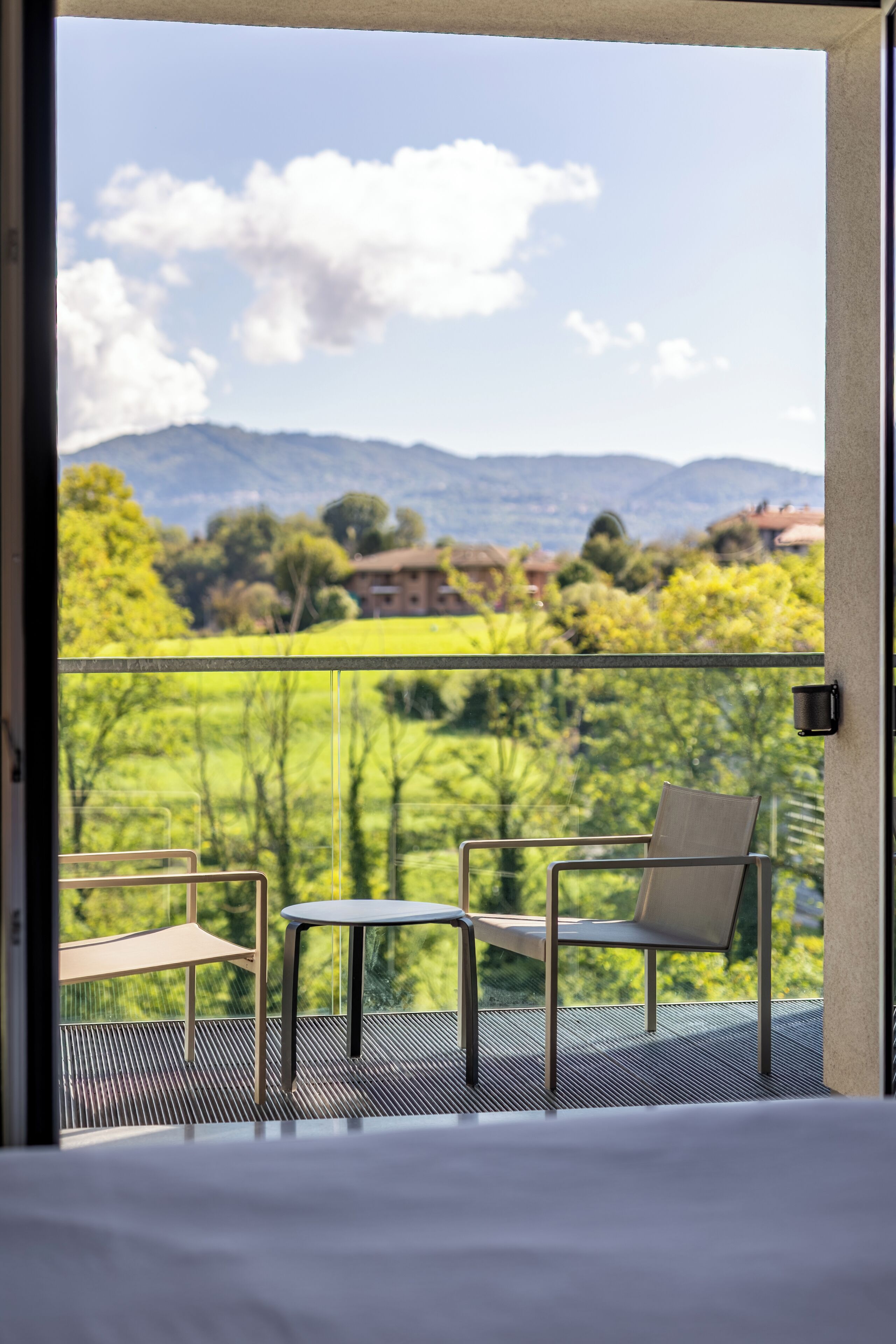 Ein Balkon mit Blick auf eine grüne Landschaft und Berge. Auf dem Balkon befinden sich ein Bett, ein Stuhl und ein kleiner Tisch.