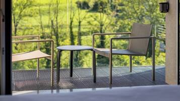 Un balcón con vistas a un paisaje verde y montañas. Se ve una cama, una silla y una mesita en el balcón.
