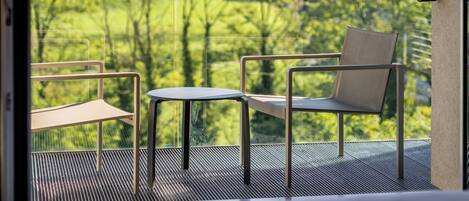 Un balcón con vistas a un paisaje verde y montañas. Se ve una cama, una silla y una mesita en el balcón.