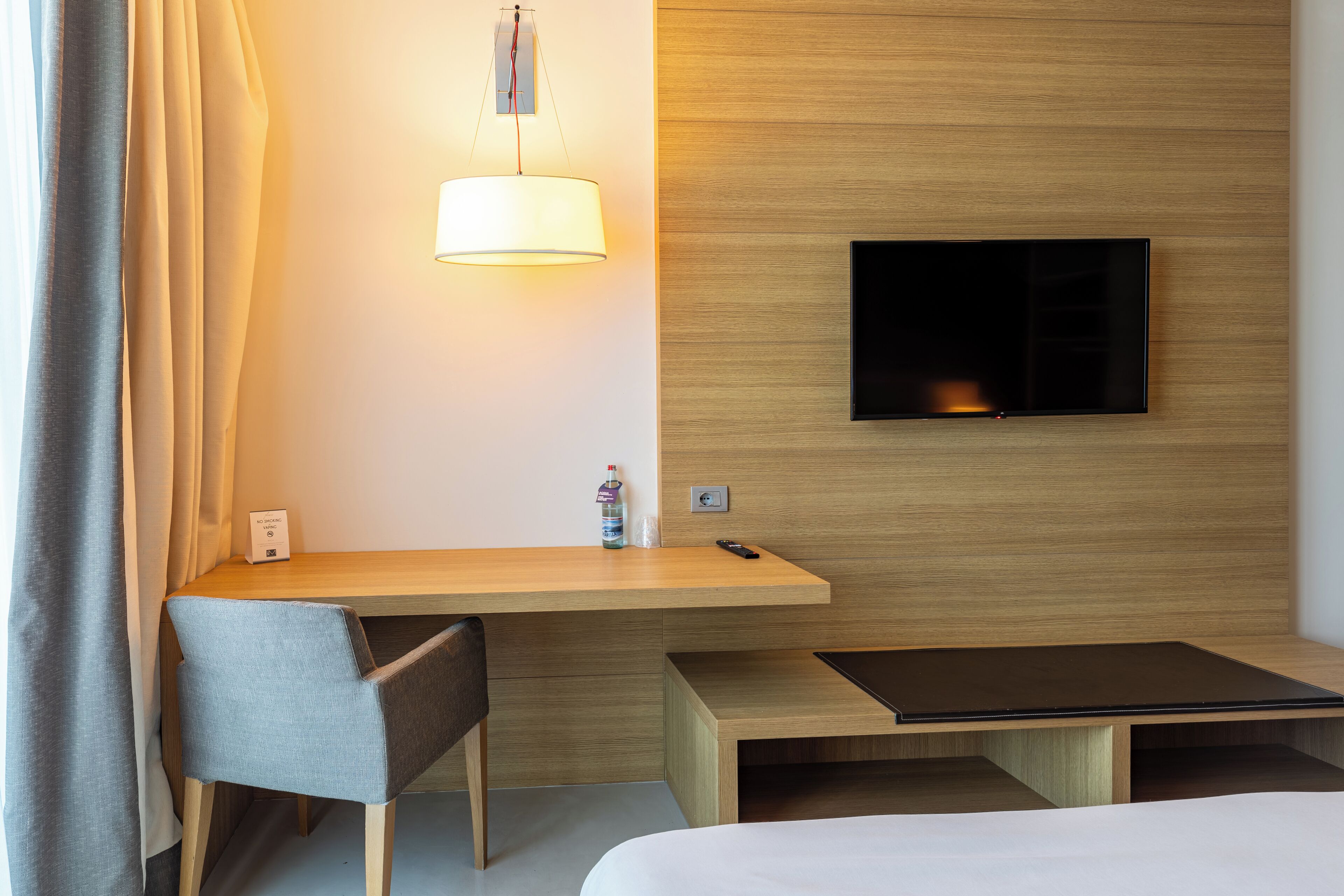 A hotel room with a wooden desk, a chair, a bed, a TV mounted on the wall, and a lamp.
