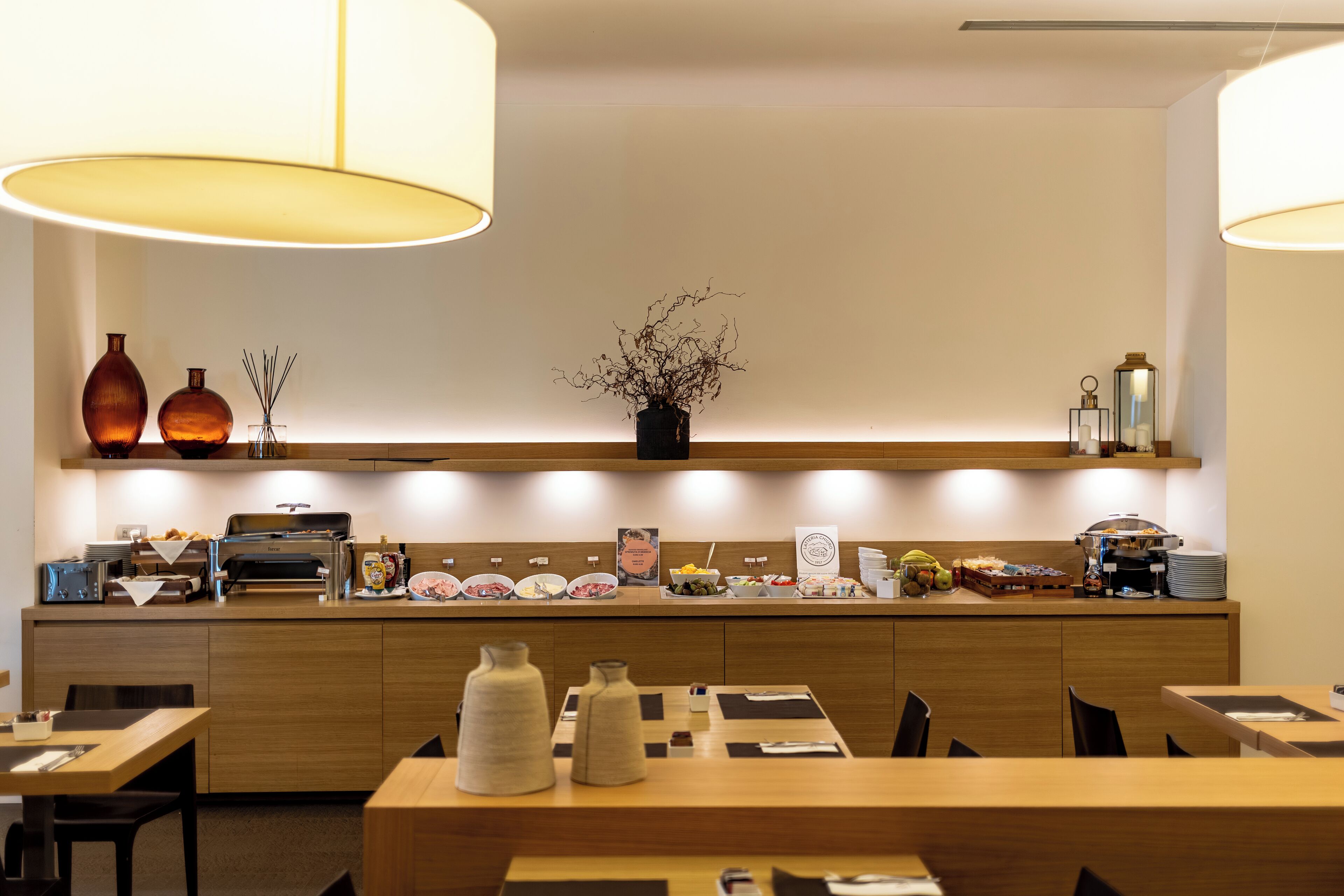 A modern dining area with wooden tables and chairs, a central island with a coffee machine, and a variety of food and drink items displayed on the counter.
