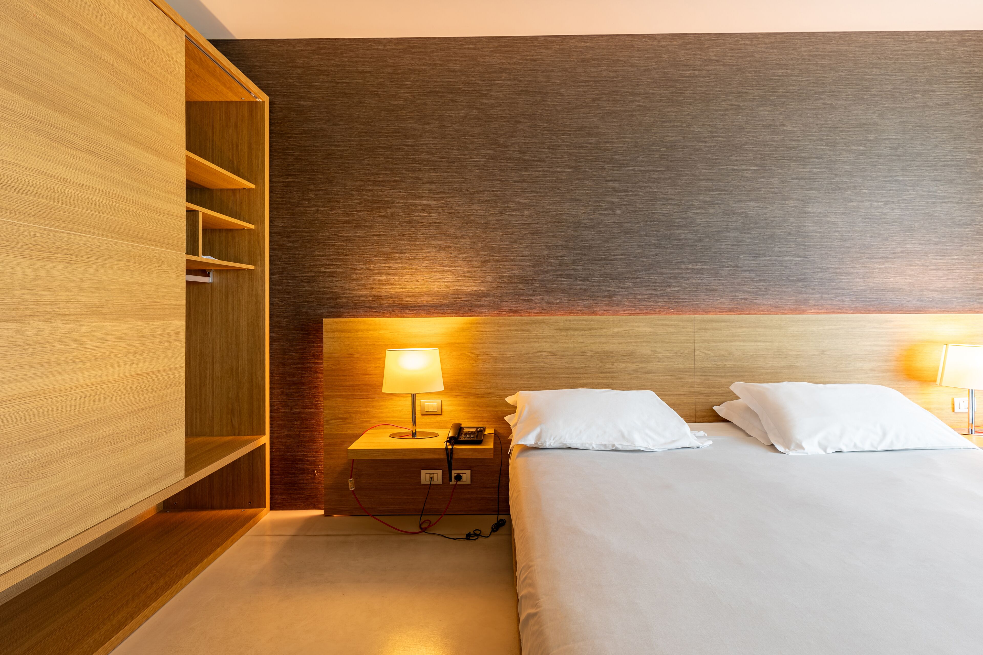 Una habitación de hotel con un armario de madera, una mesita de noche con lámpara, un teléfono y una cama con ropa blanca.