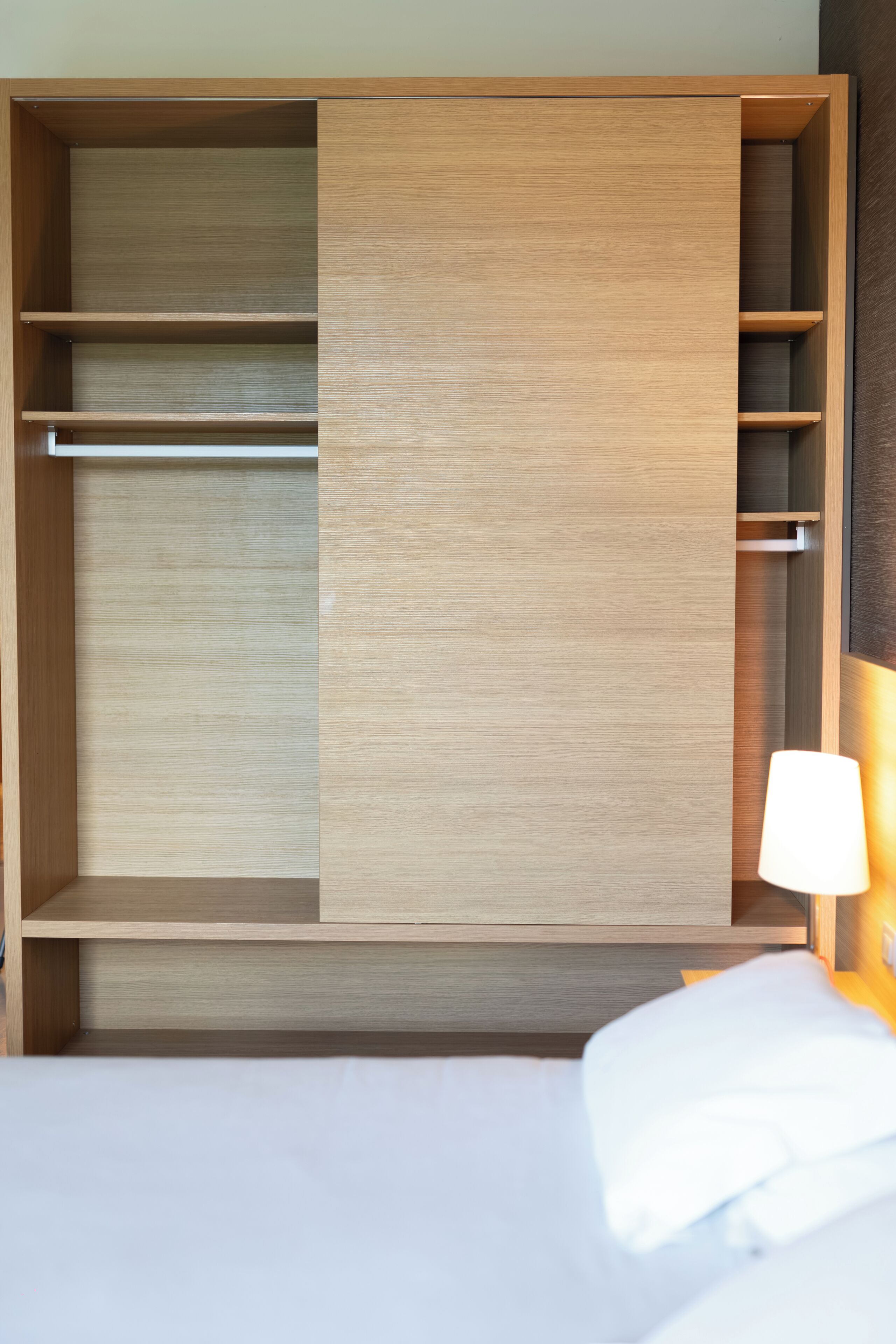Una habitación de hotel con un armario de madera, una cama con ropa blanca y una lámpara de noche.