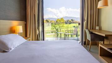 A hotel room with a bed, a desk, a chair, and a view of a balcony with a table and chairs.