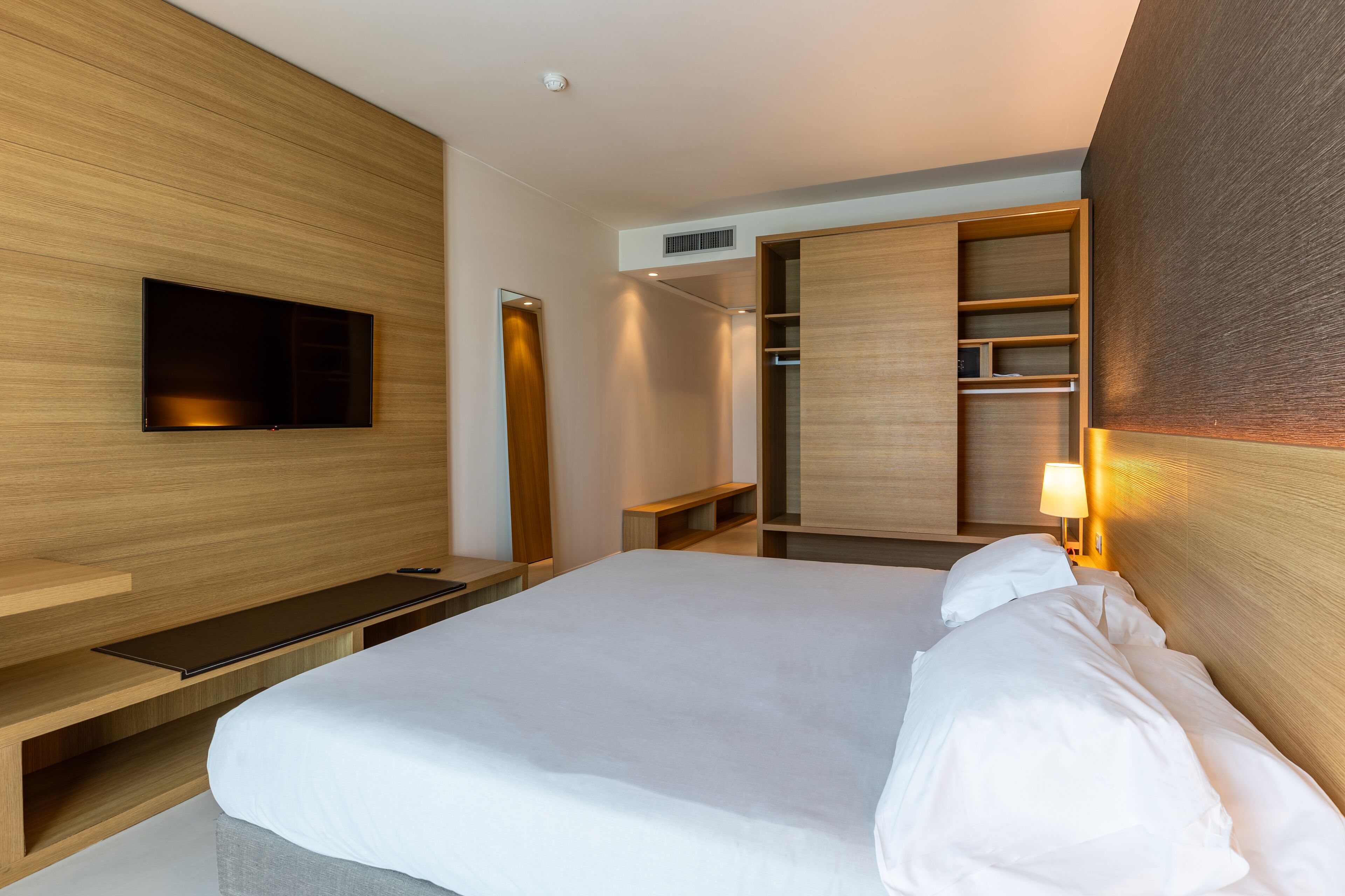 A modern hotel room with a large bed, built-in TV, wooden wardrobe, and bedside lamp.